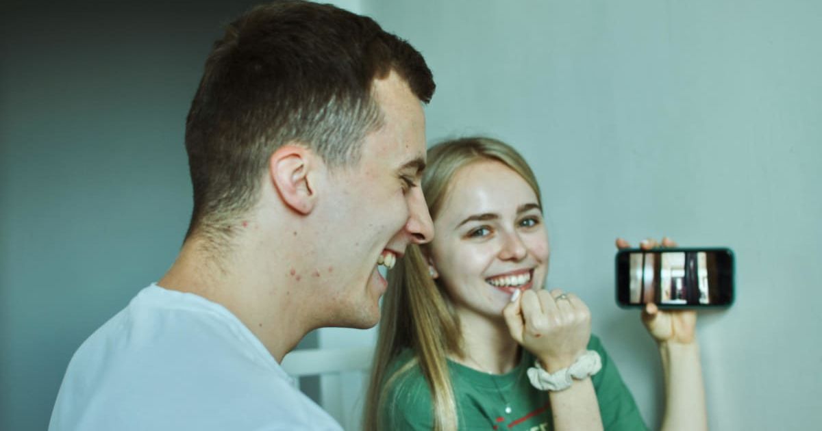 A woman is sitting with her husband, smiling and excitedly holding up her phone. Representative Cover Image Source: Pexels | Anastasiia Lopushynska