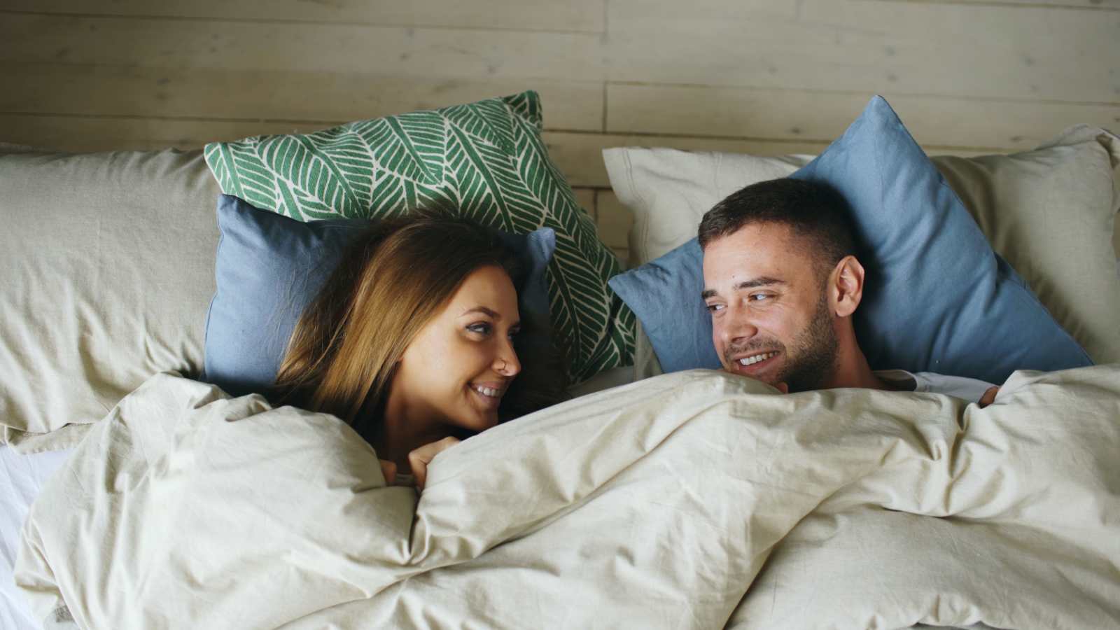 Smiling couple lying in bed with pillows and blankets, enjoying a cozy moment. (Representative image source: Pexels | Vitaly Gariev)
