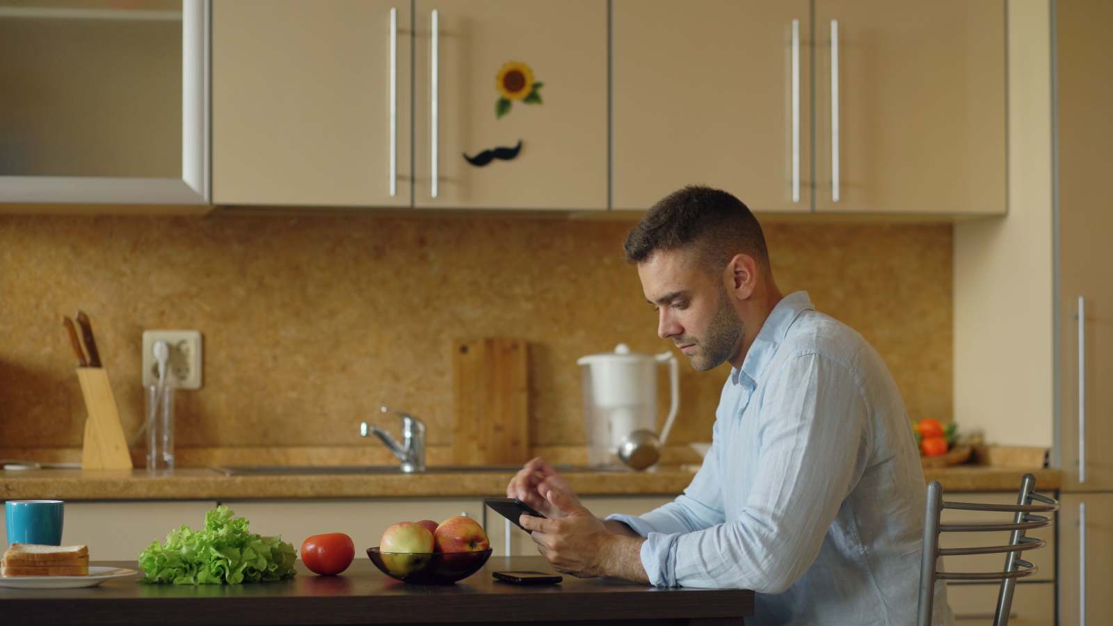 An angry man is texting in the kitchen.