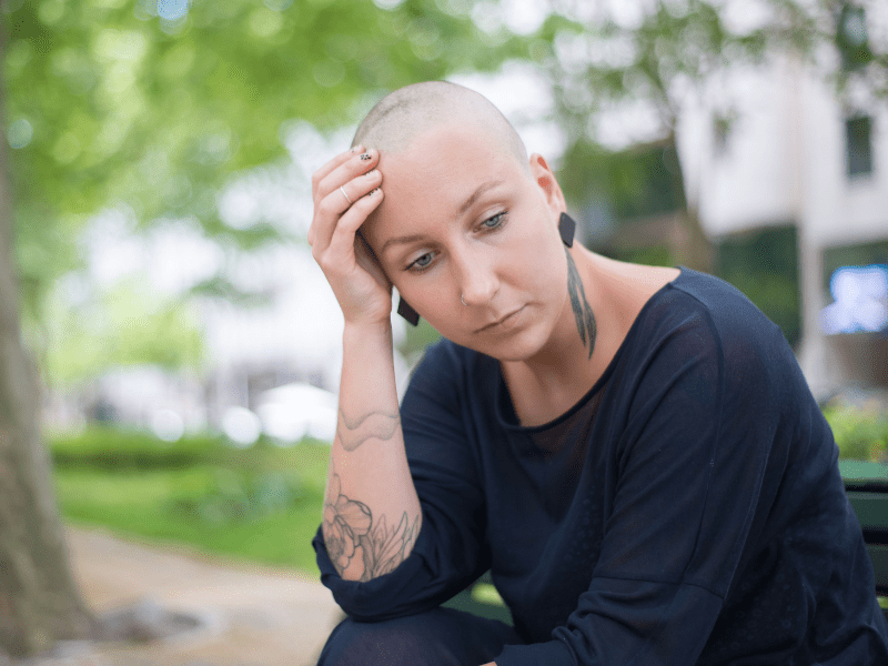 A bald woman sitting out in the park looking stressed.
