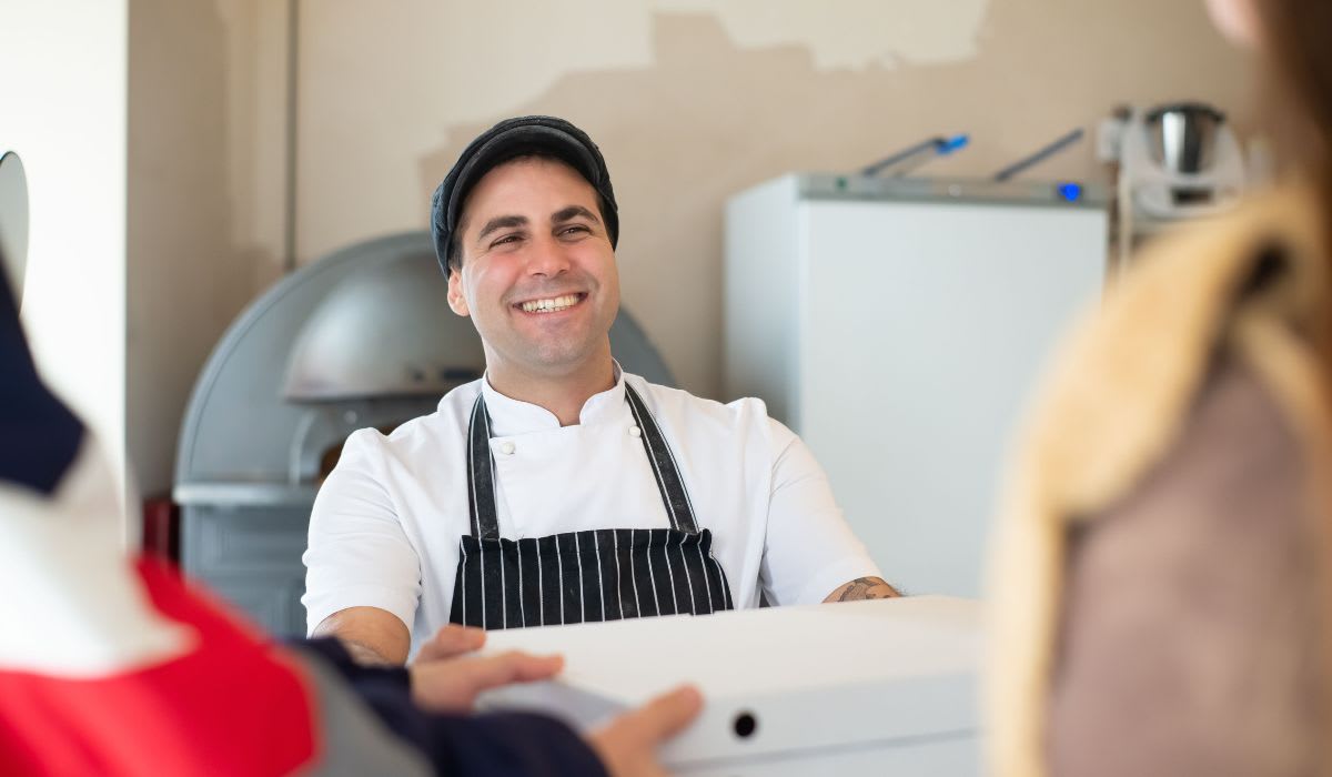 A cheerful baker in an apron hands pizza boxes to customers. (Representative Image Source: Pexels | Kampus Production)