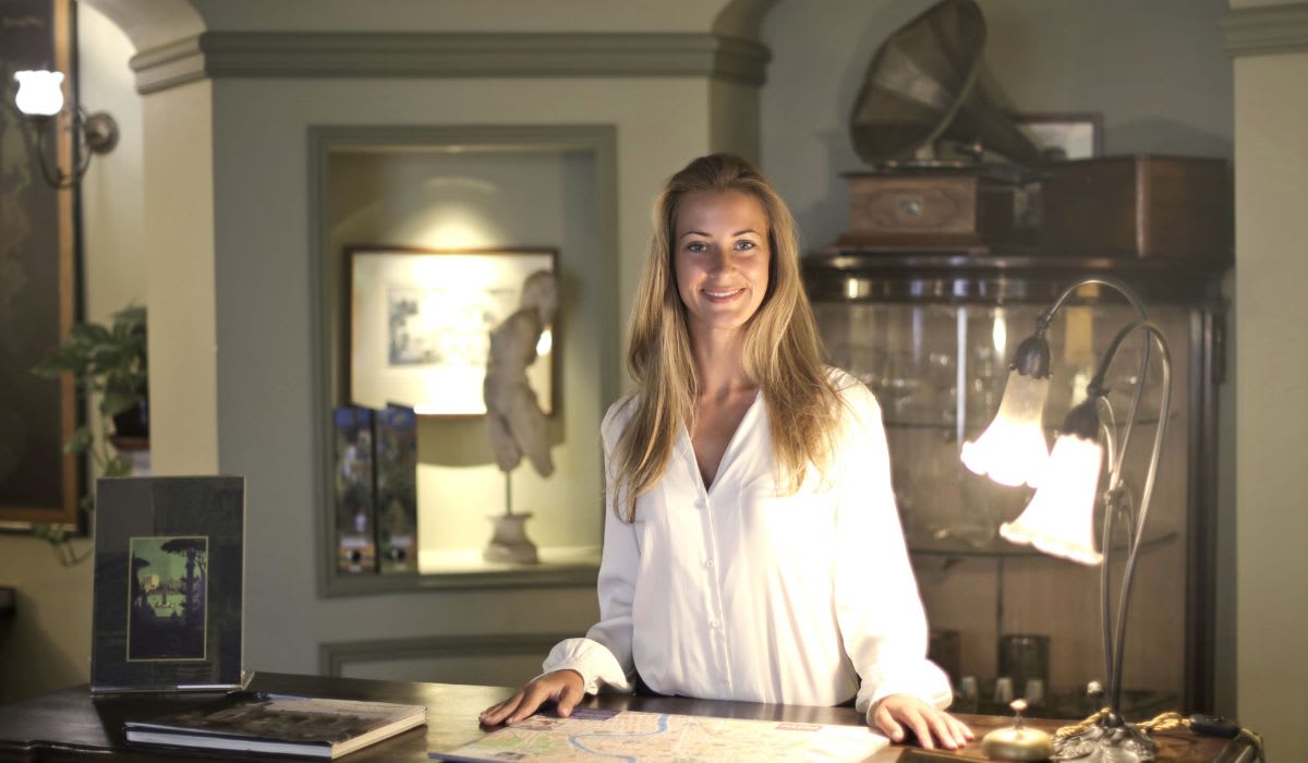 Woman smiling behind hotel reception desk. (Representative Image Source: Pexels | Andrea Piacquadio)